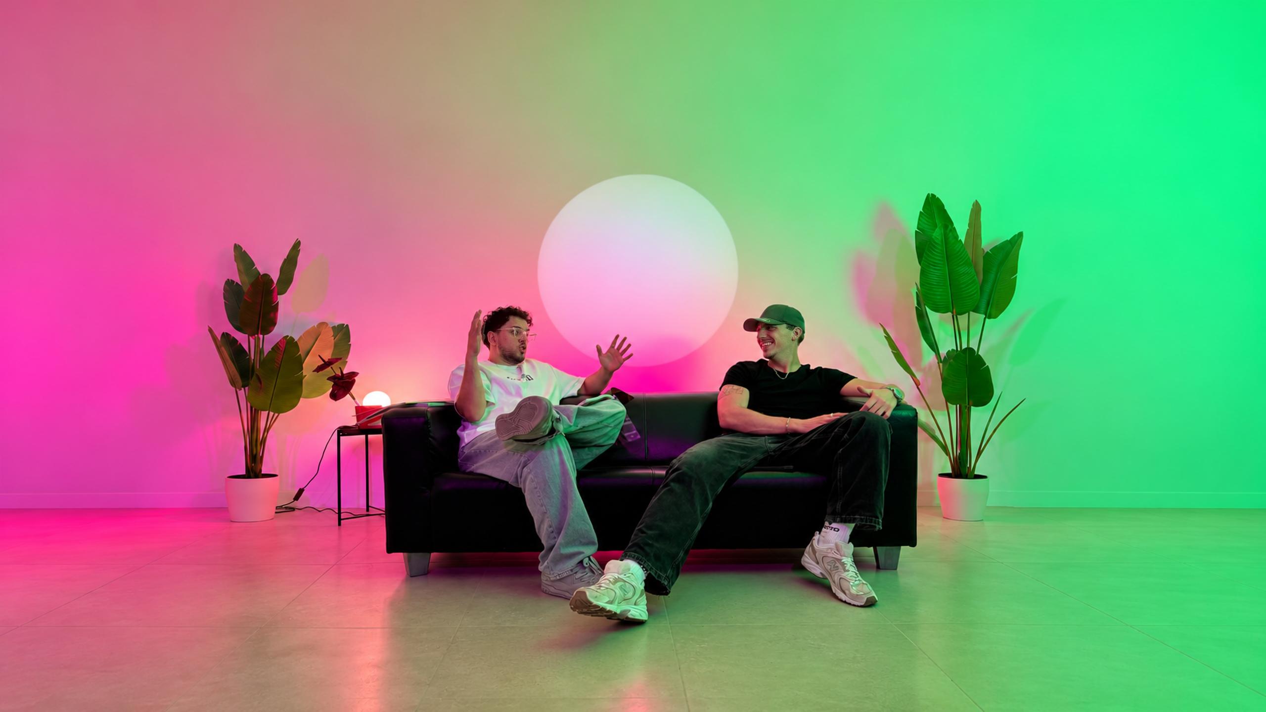 Mert und Phil auf dem schwarzen Ledersofa im pink-grün beleuchteten Studio: Mert erzählt mit beiden Händen, Phil grinst ihn an.